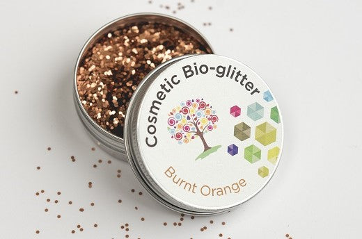 The image is  showcasing a small tin container of "Cosmetic Bio-glitter" in a shade called "Burnt Orange." The tin is open, revealing the glitter inside, which is scattered on the surface around it, emphasizing the product's texture and colour.